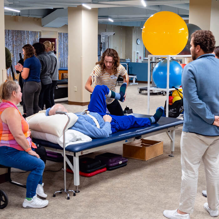 Student clinician assisting a patient with leg mobility during a therapy session in an interprofessional clinic, with faculty and peers observing.
