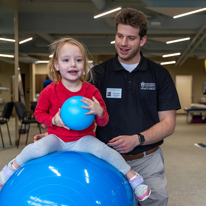 Physical therapy student supporting a young child seated on an exercise ball during a pediatric therapy activity in an interprofessional clinic.